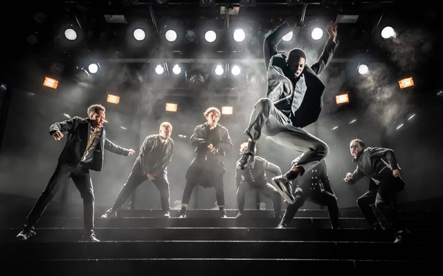 Six actors are stood on a set of stairs on stage, backlit by several stage lights. One actor is jumping down the stairs in the foreground of the image.