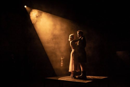 A couple dances together, illuminated by a spotlight whilst surrounded in darkness.