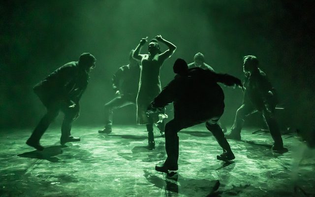 Several cast members are jumping around bathed in green light.