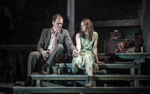 Ruth Wilson wears a green dress and sits on wooden steps near Michael Shannon. He wears a grey suit and striped tie, and looks at his hand holding Ruth’s, while she looks at him.