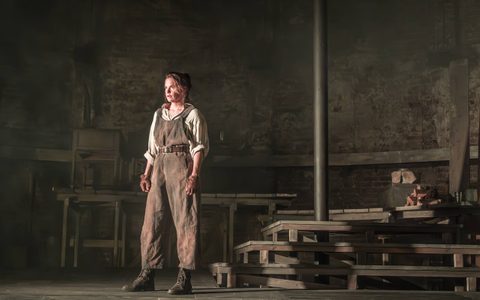 Ruth Wilson wears an off-white shirt, brown overalls and brown leather gloves. She stands on a wooden set, in front of wooden steps.
