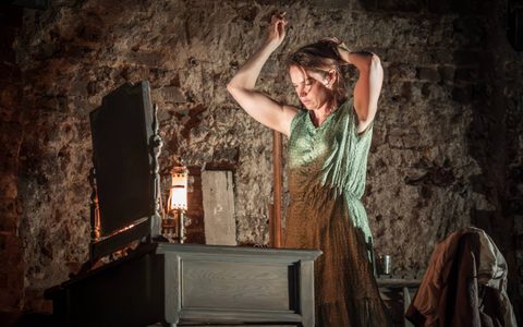 Ruth Wilson wears a green dress and stands in front of a small dressing table, putting her hair up with hair pins.