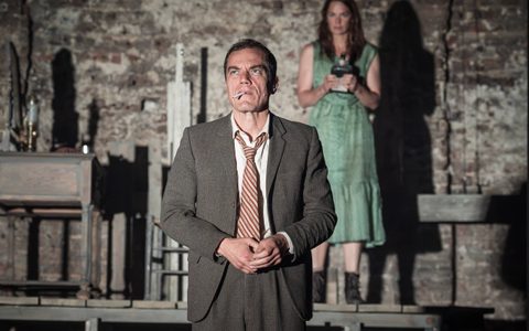 Michael Shannon wears a grey suit and striped tie. He stands with his hands together and an unlit cigarette in his mouth. Ruth Wilson stands in the background. She wears a green dress.
