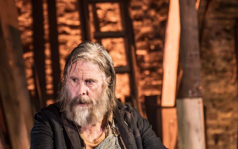 David Threlfall wears blue dungarees and a dark jacket. He sits with his hands resting on his knees.