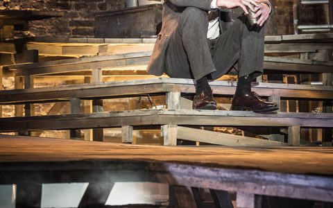 Michael Shannon wears a grey suit and sits on wooden steps. He has a neutral expression and holds a cup in front of him, resting his forearms on his knees.