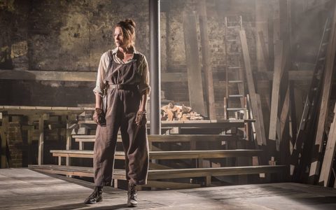 Ruth Wilson wears an off-white shirt, brown overalls and brown leather gloves. She stands on a wooden set, in front of wooden steps.