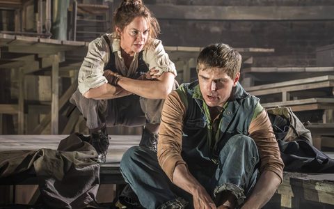 Peter Corboy wears a blue jacket and blue jeans and sits on wooden steps, lacing up his boots. Ruth Wilson crouches down behind him. She wears an off-white shirt and brown overalls, and has her arms crossed as she looks at Peter with a stern expression.