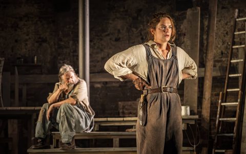 Ruth Wilson wears an off-white shirt and brown overalls and stands with her hands on her hips. She looks directly ahead with a serious expression. David Threlfall sits on wooden steps in the background.