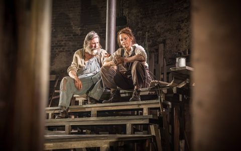 David Threlfall and Ruth Wilson sit together on wooden steps. Ruth wears an off-white shirt and brown overalls and looks down with her hands clasped. David looks at her and rests one hand on her arm.