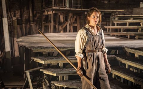 Ruth Wilson wears an off-white shirt, brown overalls and brown leather gloves. She stands on a wooden set, holding a stick.