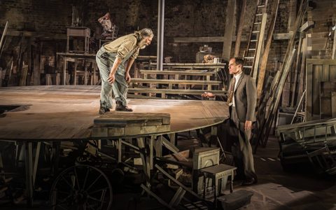 David Threlfall wears a brown shirt and blue dungarees. He stands on a circular wooden platform, leaning down to look at Michael Shannon, who wears a grey suit and stands at the edge of the stage.