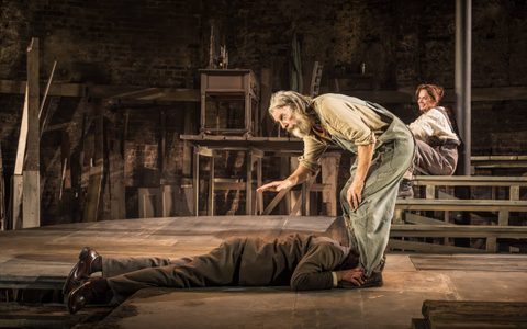 David Threlfall wears a brown shirt and blue dungarees. He stands on a circular wooden stage, leaning down to look at Michael Shannon, who wears a grey suit and lies at David’s feet. Ruth Wilson sits on wooden steps in the background and smiles.