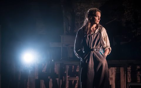Ruth wears an off-white shirt and brown overalls and stands with her hands in her pockets, looking off to the side. There is a bright white spotlight in the background.