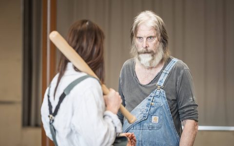 Ruth Wilson wears grey-blue dungarees and an off-white shirt. She faces away from the camera and looks at David Threlfall, who wears a grey top and blue dungarees. She holds a baseball bat over her shoulder.