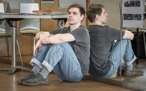 Peter Corboy wears a grey t-shirt and blue jeans. He leans against a mirror with his arms crossed over his knees.