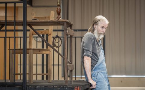 David Threlfall wears a grey top and blue dungarees. He looks off to the side as he leans back against a platform that has a table and chair on it.