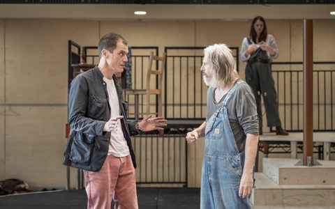 Michael Shannon wears a dark jacket and light pink jeans. He stands with his hands out in front of him, facing David Threlfall, who wears a grey top and blue dungarees. Ruth Wilson stands on a platform in the background.