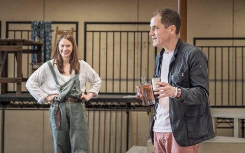 Michael Shannon wears a dark jacket and light pink jeans. He holds a glass decanter and a cup and smiles off to the side. Ruth Wilson wears grey-blue dungarees and an off-white shirt, and smiles at him with her hands on her hips.