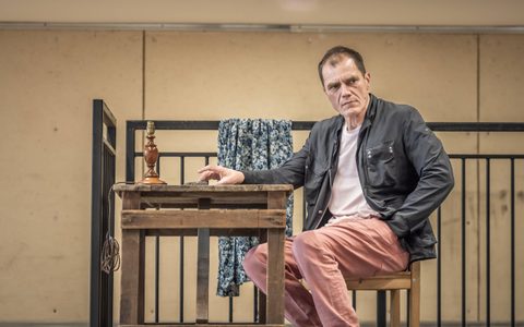 Michael Shannon wears a dark jacket and light pink jeans. He sits a wooden table and looks to the side with a serious expression.