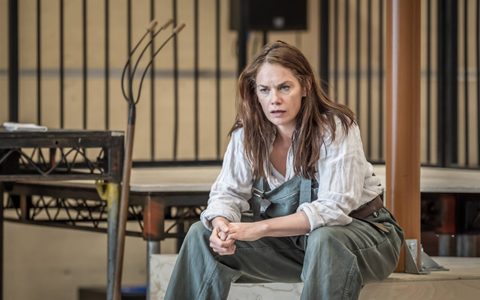 Ruth Wilson wears grey-blue dungarees and an off-white shirt. She sits wide legged with her hands clasped in front of her.