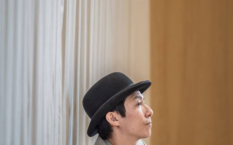 Akie Kotabe wears a black bowler hat and grey blazer. He sits leaning against a wall with one arm resting on his knee.