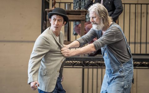 Akie Kotabe wears a black bowler hat and grey blazer. He is in a physical altercation with David Threlfall, who grabs him from his blazer.