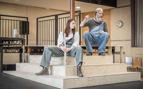 Ruth Wilson wears grey-blue dungarees and an off-white shirt, and brown boots. She looks off to the side, sitting wide legged on some steps and holding a baseball bat. David Threlfall wears a grey top and blue dungarees. He sits near Ruth.
