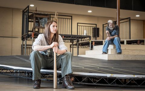 Ruth Wilson wears grey-blue dungarees and an off-white shirt, and brown boots. She sits wide legged, holding a baseball bat. David Threlfall sits on some steps in the background. He wears a grey top and blue dungarees.