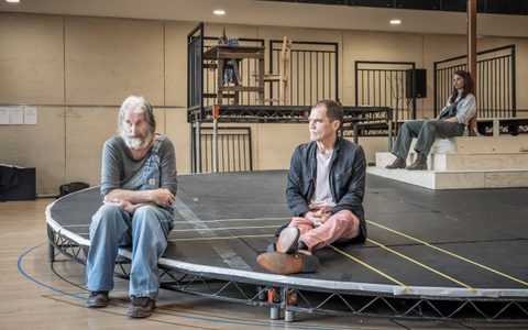 David Threlfall wears a grey top and blue dungarees. He sits with his arms crossed near Michael Shannon, who wears a dark jacket and light pink jeans. Michael looks towards David. Ruth Wilson sits on some steps in the background.