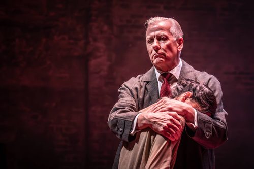 Look Back in Anger Production Photo Deka Walmsley in a white shirt with a red tie and a grey coat, stood with his arms around Ellora Torchia’s shoulders, who is leaning against his left shoulder. Her face is obscured in the crook of his elbow.