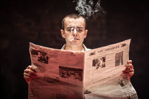 Look Back in Anger Production Photo Iwan Davies holds a newspaper open in front of him and has a cigarette in his mouth. Soft wisps of smoke billow in front of his face and above his head