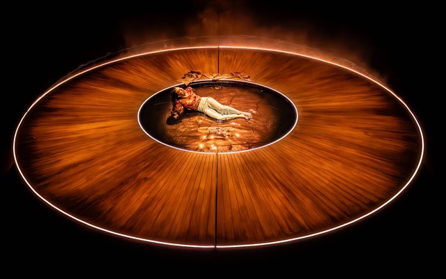 Fizz Sinclair leans back on her hands in a circular pool of water in the middle of a circular wooden stage. She is illuminated by deep orange light.