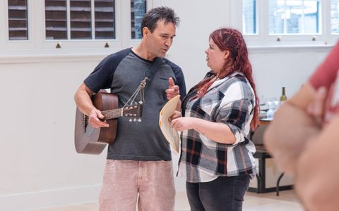 Two people stand talking in a rehearsal room. One holds a script and has a guitar around them.