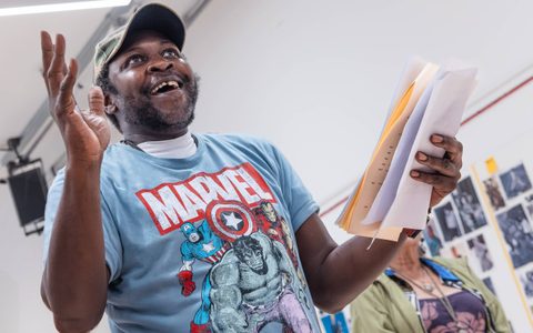 A person smiles and holds one hand up in a rehearsal room. They have a script in the other hand.