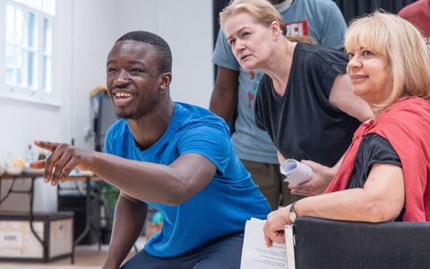 A person points and smiles in a rehearsal room. Other people look to where they are pointing.
