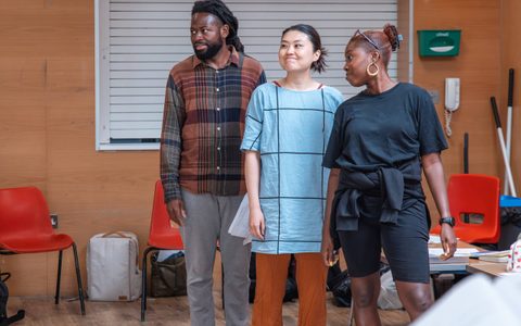 Three people stand looking off to the side in a rehearsal room.