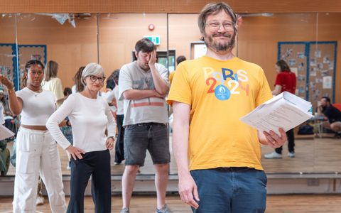 A person smiles and holds a script in a rehearsal room. Other people stand in the background.
