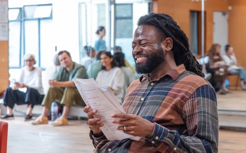 A person laughs while holding a script in a rehearsal room. Other people sit in the background against a mirror.