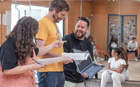 Three people smile while holding scripts in a rehearsal room.