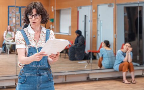 A person holds a script and looks off to the side in a rehearsal room.