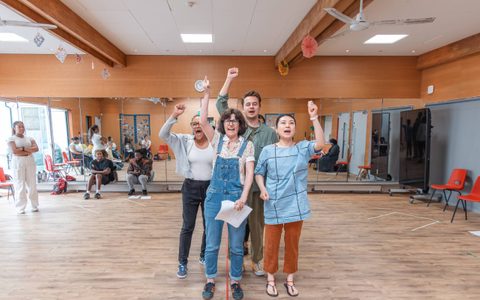A group of people raise their arms in a rehearsal room.
