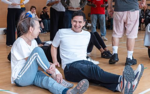 Two people sit smiling on the floor in a rehearsal room.