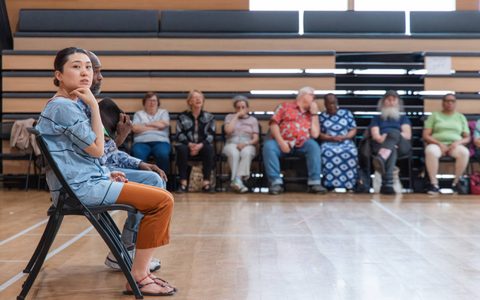 Two people sit on chairs in a rehearsal room. One looks towards the camera and a row of people sit in the background.