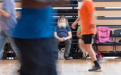 A person sits on a chair in a rehearsal room. There are blurred figures moving in the foreground.