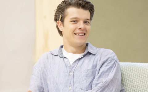 Jasper Talbot wears a blue shirt and blue jeans. He smiles, sitting on a sofa and holding a book.