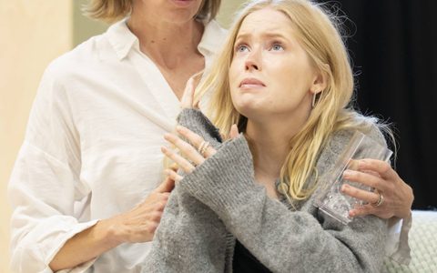 Ellie Bamber wears a grey cardigan and blue jeans. She looks up with a worried expression. Claudia Harrison sits next to her on the arm of a sofa. She has her arms around Ellie.