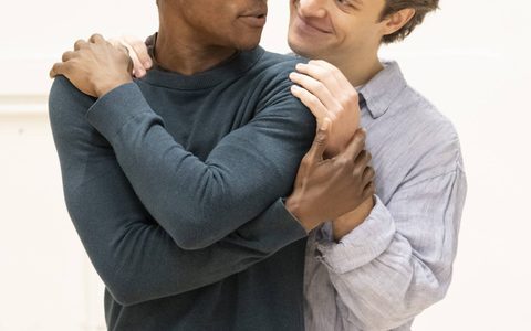 Alistair Nwachukwu looks over his shoulder at Jasper Talbot, who has his hands on Alistair’s shoulders and smiles at him, standing closely behind him.