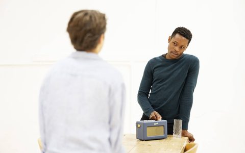 Alistair Nwachukwu wears a dark teal top and leans a wooden table with one hand. He turns the dial a radio with the other, looking at Jasper Talbot, who faces him with his back to the camera.