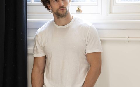 Leo Suter wears a white t-shirt and blue jeans and leans against a wall near a bright window.