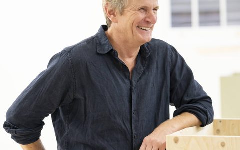 Charles Edwards stands leaning on a wooden box with arm. He is smiling.
